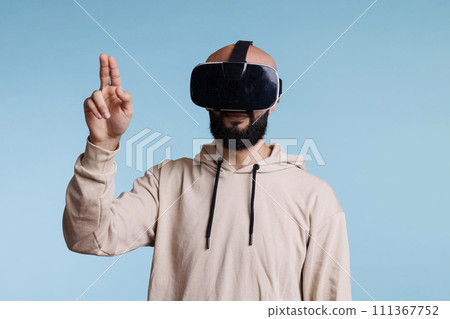 Young man in vr helmet making tap gesture with two fingers while exploring metaverse. Person in headset enjoying simulation activity and playing virtual reality games in cyberspace Young man in vr helmet making tap gesture with two fingers while exploring metaverse. Person in headset enjoying simulation activity and playing virtual reality games in cyberspace 111367752