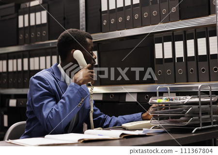 Police officer having remote conversation with inspector, discussing crime scene report using landline phone. African american detective working overhours at mysterious case in arhive room Police officer having remote conversation with inspector, discussing crime scene report using landline phone. African american detective working overhours at mysterious case in arhive room 111367804