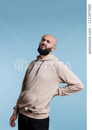 Arab man grimacing in pain while holding lower back and looking at camera. Young person wearing casual clothes suffering from chronic backache and muscles cramps portrait 111367806