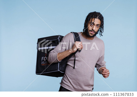 Food delivery service arab man carrying thermal bag portrait. Young deliveryman standing with backpack with restaurant take away meal and looking at camera on blue background 111367833