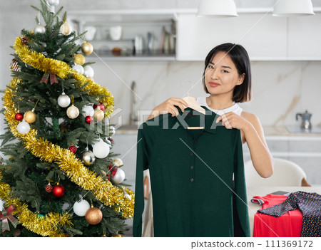 Happy young Asian girl trying to choose a dress standing next to Xmas-tree Happy young Asian girl trying to choose a dress standing next to Xmas-tree 111369172