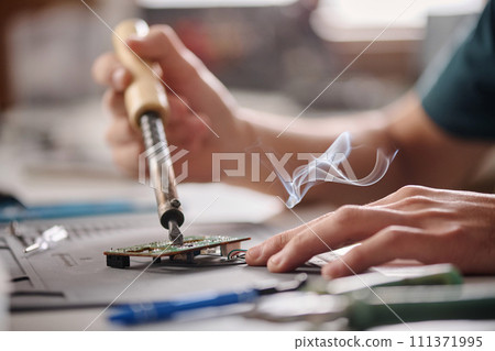 Hands of unrecognizable caucasian electronic repairman soldering mainboard sitting at workshop table 111371995