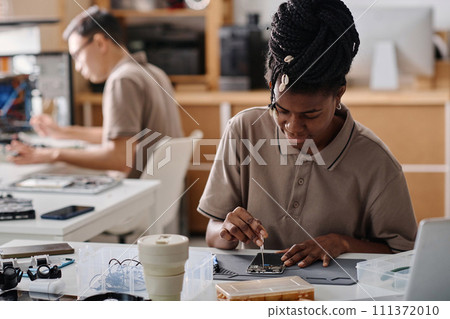 Black technician repairing broken phone with screwdriver, her male colleague working on background Black technician repairing broken phone with screwdriver, her male colleague working on background 111372010