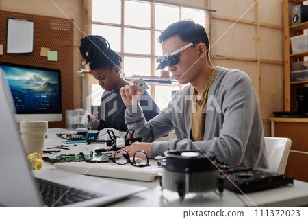 Young repairman in glasses with loupes examining CPU sitting next to his black female colleague Young repairman in glasses with loupes examining CPU sitting next to his black female colleague 111372023
