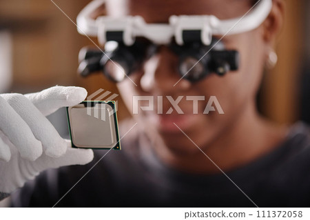 Black woman in glasses with loupes holding CPU and attentively looking at it, focus on CPU 111372058