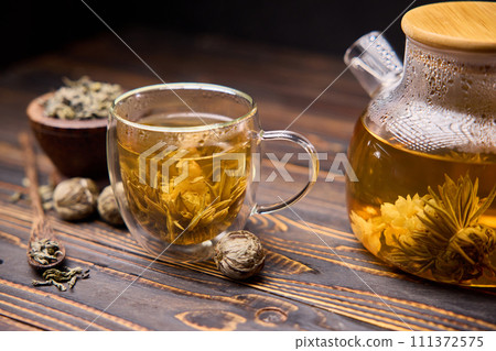 Teapot and glass cup with blooming tea flower inside on a wooden table Teapot and glass cup with blooming tea flower inside on a wooden table 111372575
