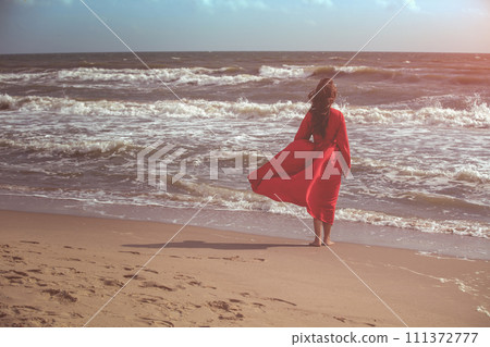 Seascape on a sunny day. Girl on the beach. A young girl in a red fluttering dress walks along the seashore. The girl looks at the sea Seascape on a sunny day. Girl on the beach. A young girl in a red fluttering dress walks along the seashore. The girl looks at the sea 111372777