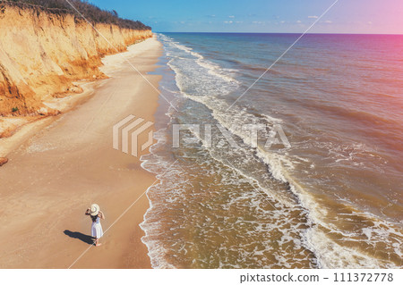 Seascape on a sunny day. Young woman in a white dress and straw hat walking on the beach. View from above 111372778