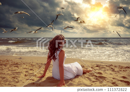 Woman relaxes on the beach listening to music on headphones and looking at the stormy sea. The woman sitting on the beach and looking at the sunset with dramatic sky Woman relaxes on the beach listening to music on headphones and looking at the stormy sea. The woman sitting on the beach and looking at the sunset with dramatic sky 111372822