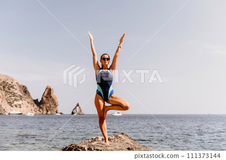 Yoga on the beach. A happy woman meditating in a yoga pose on the beach, surrounded by the ocean and rock mountains, promoting a healthy lifestyle outdoors in nature, and inspiring fitness concept. Yoga on the beach. A happy woman meditating in a yoga pose on the beach, surrounded by the ocean and rock mountains, promoting a healthy lifestyle outdoors in nature, and inspiring fitness concept. 111373144