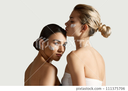 SPF face care. Brunette and blonde young girls with cream on cheeks against white studio background. Moisturizing products SPF face care. Brunette and blonde young girls with cream on cheeks against white studio background. Moisturizing products 111375706