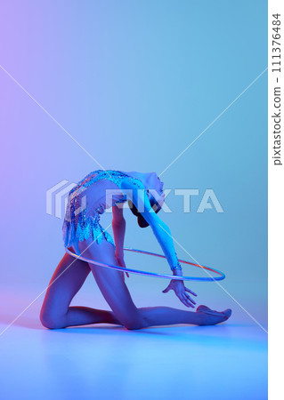 Flexible, talented teen girl, rhythmic gymnast in sparkling bodysuit performing with hoop against gradient studio background in neon light Flexible, talented teen girl, rhythmic gymnast in sparkling bodysuit performing with hoop against gradient studio background in neon light 111376484