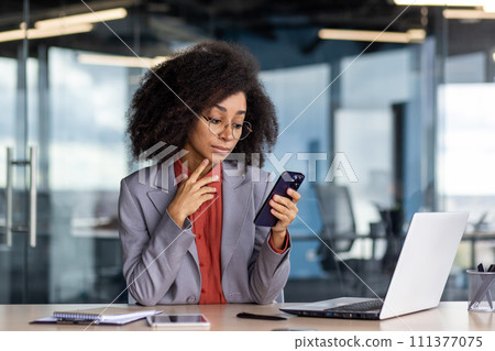 Focused woman with curly hair using smartphone while working in a contemporary office setting, showcasing multitasking and efficiency. 111377075