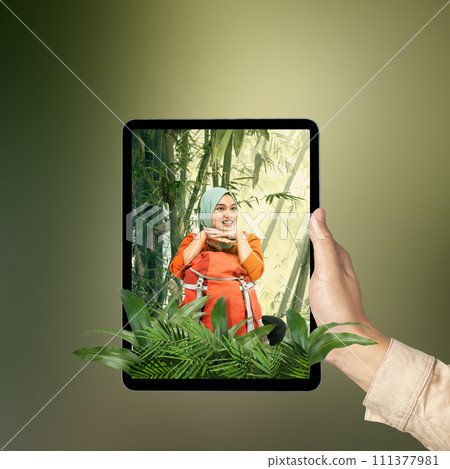 A human hand holding tablet with a screen view of an Asian girl in a scarf sitting with a backpack in the rainforest with trees and sunlight 111377981