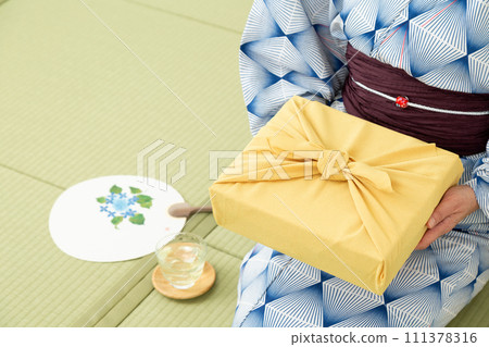 A middle-aged woman in a yukata sitting on a tatami mat in a Japanese-style room and holding a wrapping paper as a summer gift. 111378316