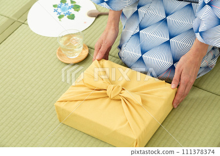 A middle-aged woman in a yukata sitting on a tatami mat in a Japanese-style room and holding a wrapping paper as a summer gift. 111378374
