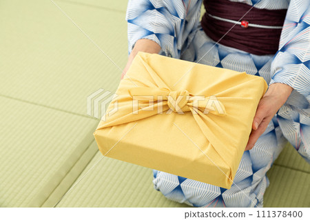 一位身穿浴衣的中年婦女坐在日式房間的榻榻米上,手裡拿著一份包裝紙,作為夏日禮物。 一位身穿浴衣的中年婦女坐在日式房間的榻榻米上,手裡拿著一份包裝紙,作為夏日禮物。 111378400