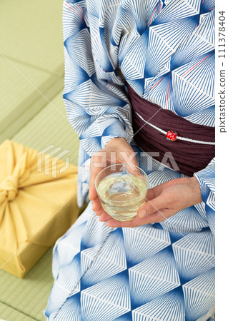 一位穿著浴衣的中年婦女坐在日式房間的榻榻米上,喝著冷茶,拿著年中禮物的包裝布 一位穿著浴衣的中年婦女坐在日式房間的榻榻米上,喝著冷茶,拿著年中禮物的包裝布 111378404