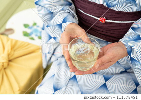 A middle-aged woman in a yukata sitting on a tatami mat in a Japanese-style room drinking cold tea and a wrapping cloth for a midyear gift 111378451