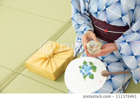 A middle-aged woman in a yukata sitting on a tatami mat in a Japanese-style room drinking cold tea and a wrapping cloth for a midyear gift 111378461