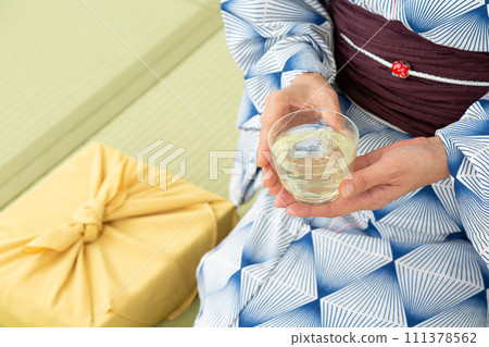 A middle-aged woman in a yukata sitting on a tatami mat in a Japanese-style room drinking cold tea and a wrapping cloth for a midyear gift 111378562