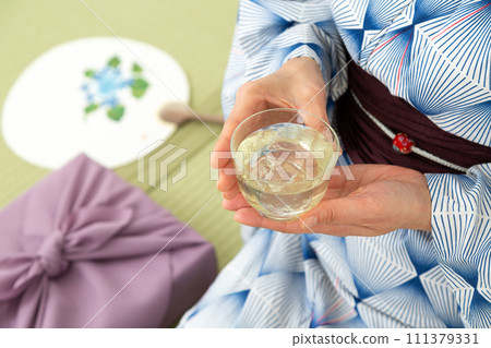 A middle-aged woman in a yukata sitting on a tatami mat in a Japanese-style room drinking cold tea and a wrapping cloth for a midyear gift 111379331