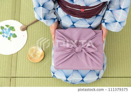 A middle-aged woman in a yukata sitting on a tatami mat in a Japanese-style room and holding a wrapping paper as a summer gift. A middle-aged woman in a yukata sitting on a tatami mat in a Japanese-style room and holding a wrapping paper as a summer gift. 111379375