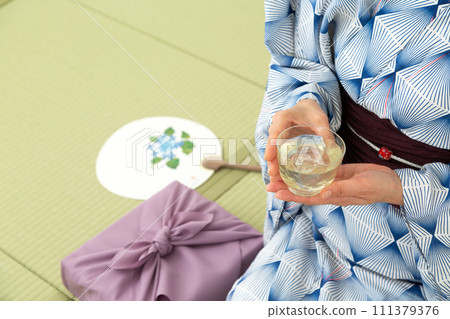 A middle-aged woman in a yukata sitting on a tatami mat in a Japanese-style room drinking cold tea and a wrapping cloth for a midyear gift A middle-aged woman in a yukata sitting on a tatami mat in a Japanese-style room drinking cold tea and a wrapping cloth for a midyear gift 111379376