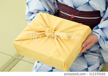 A middle-aged woman in a yukata sitting on a tatami mat in a Japanese-style room and holding a wrapping paper as a summer gift. A middle-aged woman in a yukata sitting on a tatami mat in a Japanese-style room and holding a wrapping paper as a summer gift. 111379383