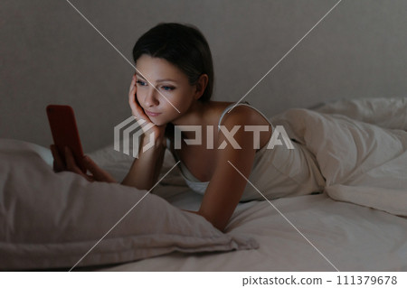 Young woman holding smartphone in her hand and watching series in bed at night 111379678