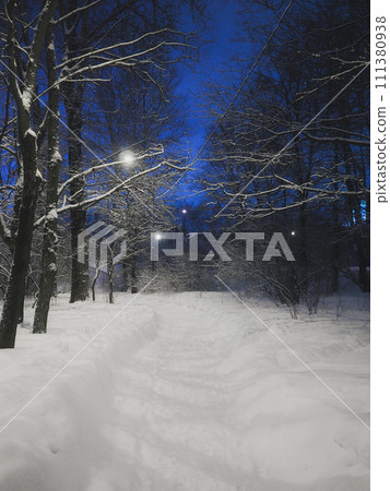 A snow-covered alley of a city park illuminated by lanterns.  111380938