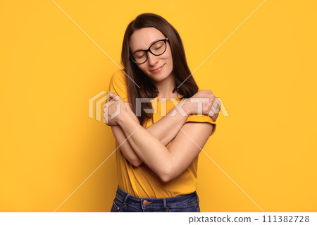 Pretty caucasian woman in eyeglasses hugging herself isolated on yellow studio wall 111382728