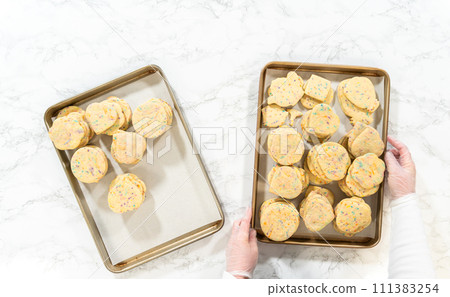 Flat lay. Large quantity of sprinkle-filled sugar cookies are neatly stacked on a baking tray, awaiting their packaging into the rustic brown paper boxes. 111383254