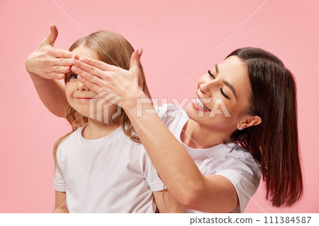 Smiling beautiful young woman, mother covering her little daughter's eyes, surprising and playing with kid against pink studio background Smiling beautiful young woman, mother covering her little daughter's eyes, surprising and playing with kid against pink studio background 111384587