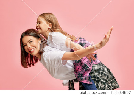 Joyful, happy, smiling mother giving her little daughter piggyback ride, playing against pink studio background 111384607
