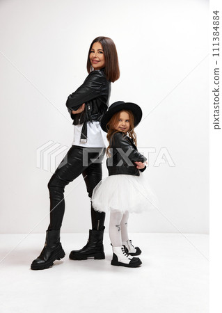 Portrait of happy mother posing with little kid, daughter in same style black and white outfit against white studio background. Family bonding 111384884