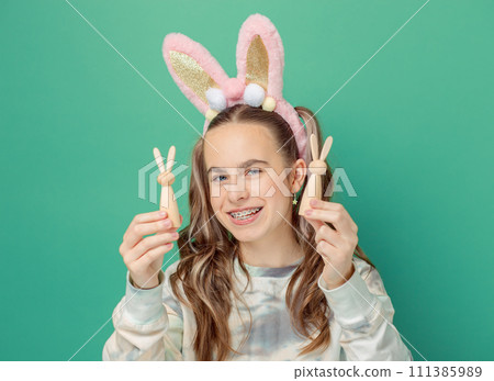 Girl with joyful smile, wearing glittery pink bunny ears holding wooden bunny figures, showcases her braces Girl with joyful smile, wearing glittery pink bunny ears holding wooden bunny figures, showcases her braces 111385989