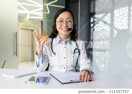Smiling Asian female doctor in clinic office offers telehealth consultation, equipped with headset for remote medical assistance to patients. 111387707