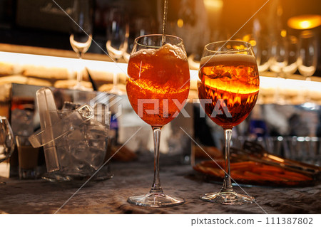 Two glasses of cocktail Aperol spritz on bar counter. Making typical alcoholic italian beverage, aperitif made with Prosecco sparkling white wine, ice and fresh orange slices. Mixing orange liquid. Two glasses of cocktail Aperol spritz on bar counter. Making typical alcoholic italian beverage, aperitif made with Prosecco sparkling white wine, ice and fresh orange slices. Mixing orange liquid. 111387802