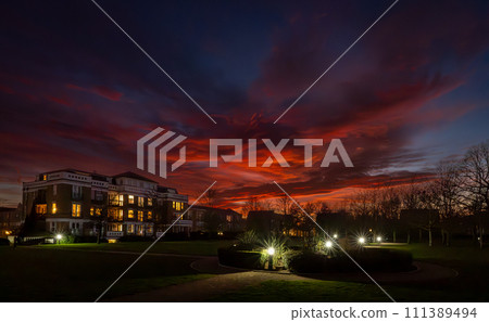Park with lights at sunset with a fiery red sky. Night view in landscape orientation with buildings and trees behind. 111389494