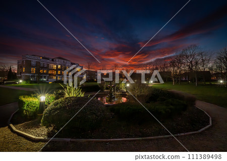 Fountain and pond in a park at sunset with a red sky. Wide angle night view in landscape orientation with buildings and trees behind. Fountain and pond in a park at sunset with a red sky. Wide angle night view in landscape orientation with buildings and trees behind. 111389498