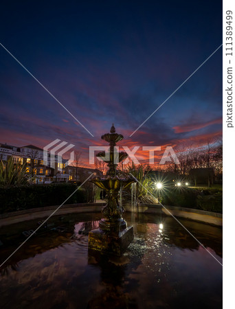 Fountain and pond in a park at sunset with a pink sky. Night view in portrait orientation with buildings and trees behind. Fountain and pond in a park at sunset with a pink sky. Night view in portrait orientation with buildings and trees behind. 111389499