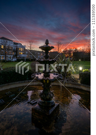 Fountain and pond in a park at sunset with a pink sky. Dusk view in portrait orientation with buildings and trees behind. 111389500