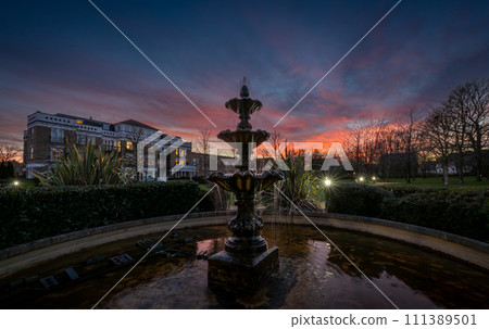 Fountain and pond in a park at sunset with a pink sky. Dusk view in landscape orientation with buildings and trees behind. 111389501