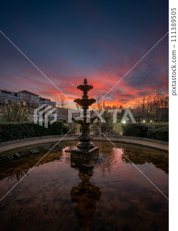 Fountain and pond in a park at sunset with a pink sky. Evening view in portrait orientation with buildings and trees behind. 111389505