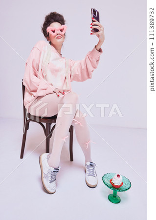Young pretty woman sitting on chair in pink comfortable clothes and sunglasses, taking selfie with mobile phone. Social media campaign 111389732