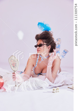 Beautiful young woman with feather head accessories and sunglasses lying on bed in vintage lingerie and looking on antique mirror 111389754