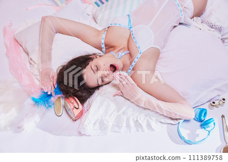 Pretty young woman in vintage sleepwear, yawning, lying on bed against pink pastel background. Wellness and focus on quality of sleep 111389758