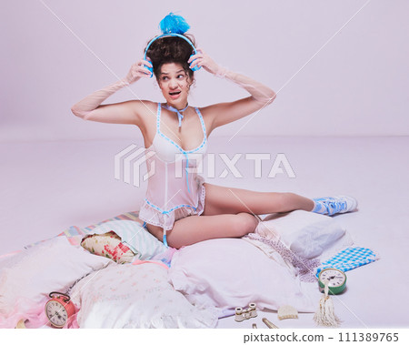 Too big hairstyle. Young woman sitting in elegant lingerie on bed, trying to wear headphones against pastel pink background. Too big hairstyle. Young woman sitting in elegant lingerie on bed, trying to wear headphones against pastel pink background. 111389765