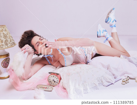 Tired woman in vintage sleepwear, yawning, lying on bed against pink pastel background. Relaxation and meditation app visual 111389785